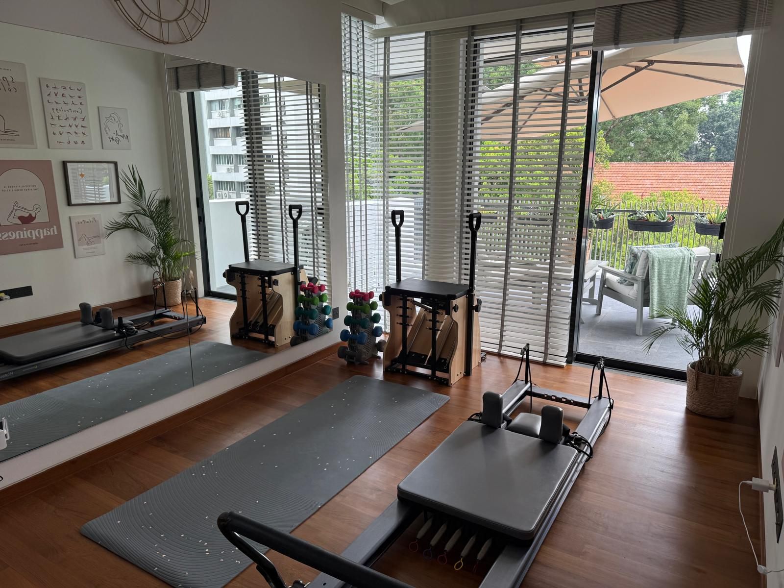 The studio: a reformer, chair and mat on a wooden floor, beside tall windows and a balcony with greenery.